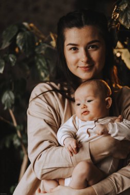 Genç esmer anne ve yeni doğmuş bebeği kollarında, karanlık bir odada, pencerenin kenarında.
