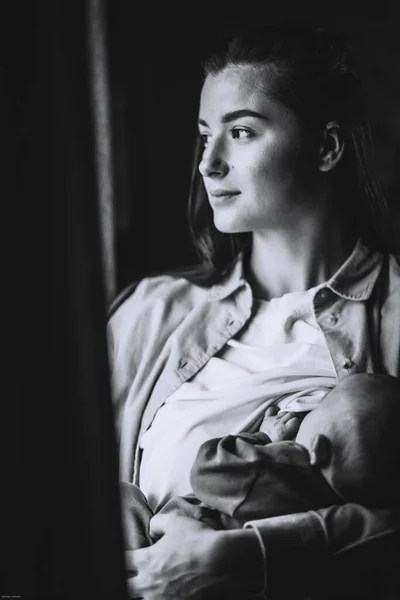 若いブルネットの母親と彼女の腕の中の新生児窓のそばの暗い部屋の家の写真 — ストック写真