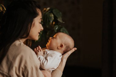 Sevgi dolu anne, yeni doğmuş bebeğini akşam evde taşıyor. Bebek kucağında mutlu bir annenin samimi portresi. Anne 3 aylık kızına sarılıyor.