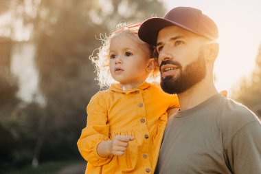 Havalı sakallı baba, küçük kıvırcık kızını tutuyor.