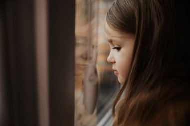 Güzel küçük kız gülümser ve pencereden dışarı bakar. Çocuk pencereden dışarı bakıyor. Genç bir kız pencereden dışarı bakıyor. Pencere kenarında neşeli bir çocuğun portresi..