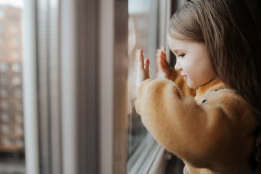 Pencerenin kenarındaki küçük bir kız yüksek binadaki daireden dışarı bakıyor.
