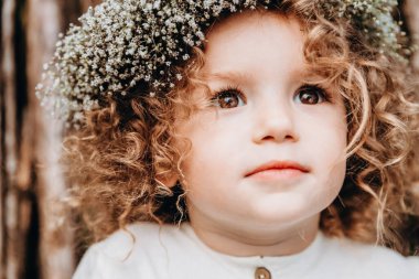 3 yaşında güzel, kıvırcık saçlı ve başında çelenk olan bir kız çocuğu doğadaki bir fotoğraf için poz veriyor.