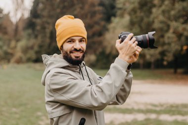 Elinde fotoğraf makinesi olan şık sakallı bir fotoğrafçı sonbahar parkında fotoğraf çekiyor.