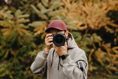 Elinde fotoğraf makinesi olan şık sakallı bir fotoğrafçı sonbahar parkında fotoğraf çekiyor.