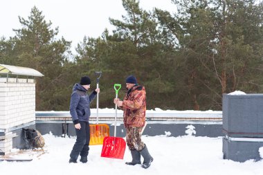 Binanın çatısında kar temizleme. Arka planda, çam ağaçlarının tepeleri ve gri gökyüzü. Sıcak kış kıyafetleri içinde kar kürekleri olan bir adam ve bir adam..