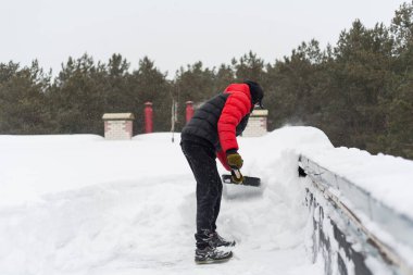 İlkbaharda çatıdan kar kaldırılacak. Güçlü kar küreği ve büyük kar yığınları. Arka planda, çam ağaçlarının tepeleri ve bulutlu bir gökyüzü..