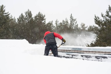 İlkbaharda çatıdan kar kaldırılacak. Güçlü kar küreği ve büyük kar yığınları. Arka planda, çam ağaçlarının tepeleri ve bulutlu bir gökyüzü..