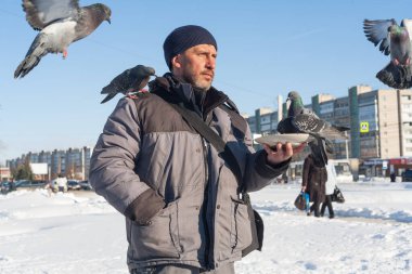 Bir adam elleriyle güvercinleri besler. İnsanlar şehirde kışın vahşi kuşlara bakarlar. Arka planda, karla kaplı, soğuk bir şehir ve mavi bir gökyüzü.