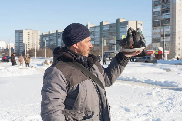 A man feeds pigeons with his hands. People take care of wild birds in winter in the city. In the background, a snow-covered, cold city, and a blue sky