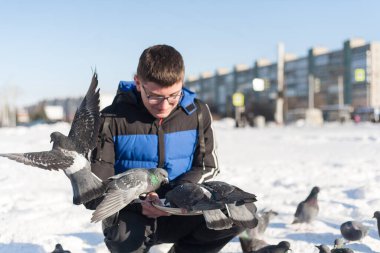 Gözlüklü ve kafasında gençlik saç stili olan genç bir adam, elleri kışın şehrin arka planında güvercinleri besliyor..