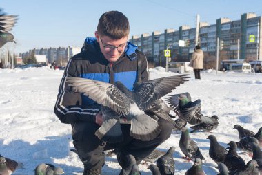 Gözlüklü ve kafasında gençlik saç stili olan genç bir adam, elleri kışın şehrin arka planında güvercinleri besliyor..