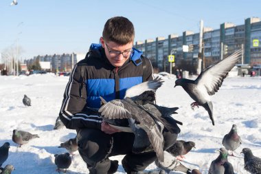 Gözlüklü ve kafasında gençlik saç stili olan genç bir adam, elleri kışın şehrin arka planında güvercinleri besliyor..