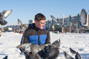 Gözlüklü ve kafasında gençlik saç stili olan genç bir adam, elleri kışın şehrin arka planında güvercinleri besliyor..