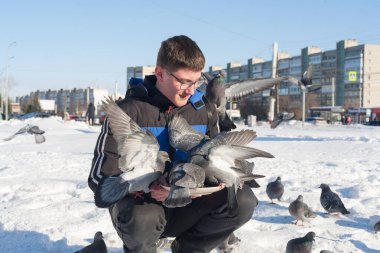 Gözlüklü ve kafasında gençlik saç stili olan genç bir adam, elleri kışın şehrin arka planında güvercinleri besliyor..