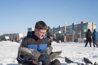 Gözlüklü ve kafasında gençlik saç stili olan genç bir adam, elleri kışın şehrin arka planında güvercinleri besliyor..