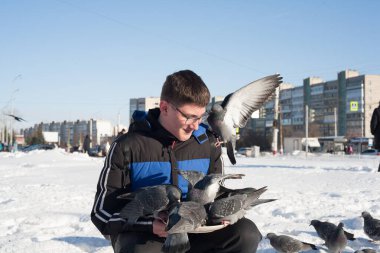Gözlüklü ve kafasında gençlik saç stili olan genç bir adam, elleri kışın şehrin arka planında güvercinleri besliyor..