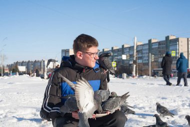 Gözlüklü ve kafasında gençlik saç stili olan genç bir adam, elleri kışın şehrin arka planında güvercinleri besliyor..