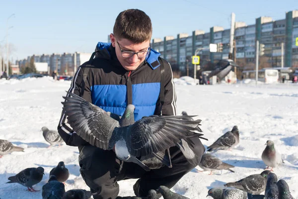 Gözlüklü ve kafasında gençlik saç stili olan genç bir adam, elleri kışın şehrin arka planında güvercinleri besliyor..