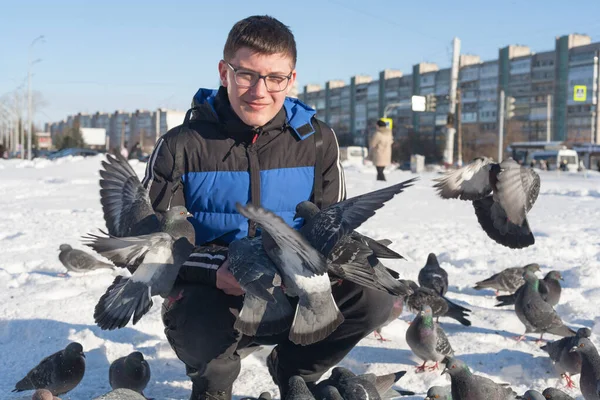 Gözlüklü ve kafasında gençlik saç stili olan genç bir adam, elleri kışın şehrin arka planında güvercinleri besliyor..