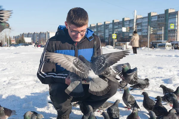 Gözlüklü ve kafasında gençlik saç stili olan genç bir adam, elleri kışın şehrin arka planında güvercinleri besliyor..