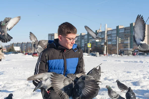 Gözlüklü ve kafasında gençlik saç stili olan genç bir adam, elleri kışın şehrin arka planında güvercinleri besliyor..