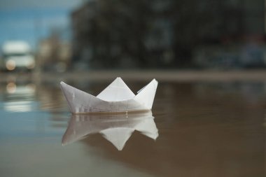 Kağıttan bir tekne su birikintisinde yüzer, bir araba arka planda gider.