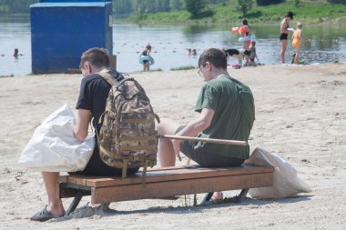 Gençler şehir plajında çöp toplamakla meşgul. Sıcak yazda gölde temizlik yapmak. Temizlik beyaz çöp torbasına çivi çakılmış bir sopayla yapılır..