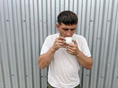 Beyaz tişörtlü bir adamın elinde canlandırıcı bir fincan çay yuvarlak bir duvarın arka planında, küçük beyaz bir bardaktan canlandırıcı bir çay içen bir adam..