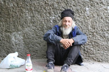 Yaşlı bir adam beton bir duvara yaslanmış asfaltta oturuyor. Uzun gri saçlı ve üzgün gözlü bir adam dünyaya bakıyor. Yaşlı adamın ayaklarının dibinde bir şişe süt ve bir torba yiyecek var. Giysileri eski ama hâlâ güçlü. Rusya