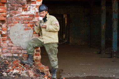 Bir inşaat işçisi eski bir fabrikanın arka planında kahve içer. Koruyucu kaskın güneşliğinde yazıt yapımcısı siyahtır. İş elbiseli bir adamın ellerinde kahve olan beyaz bir kupa..