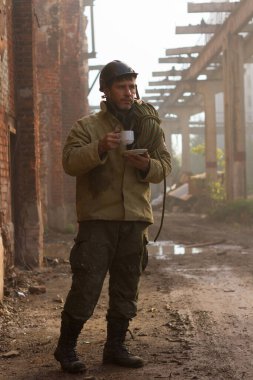 Bir inşaat işçisi eski bir fabrikanın arka planında kahve içer. Koruyucu kaskın güneşliğinde yazıt yapımcısı siyahtır. İş elbiseli bir adamın ellerinde kahve olan beyaz bir kupa..