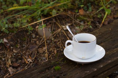 Beyaz çay tabağında kaşığı olan beyaz bir kupa yeşil çimenlerin arka planında eski bir tahta üzerinde duruyor..
