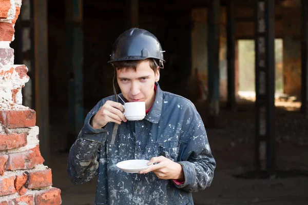 A construction worker drinks coffee against the background of an old factory. On the visor of the protective helmet, the inscription builder is black. A white cup with coffee in the hands of a man in work clothes.