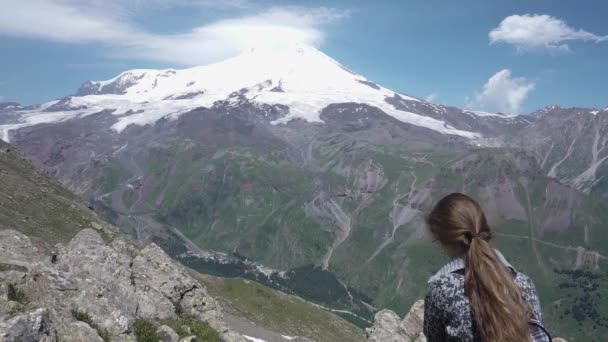 Een meisje zit op een steen op de helling van de berg Cheget en kijkt naar de vulkaan Elbrus. Kabardino-Balkaria Republiek. Rusland — Stockvideo