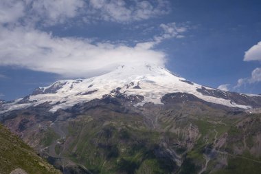 Volkan Elbrus 'un zirveleri güneşli havada bulutlarla örtülüdür. Cheget Dağı 'nın güneydoğusundaki manzara manzarası. Kabardino-Balkaria bölgesi, Rusya.