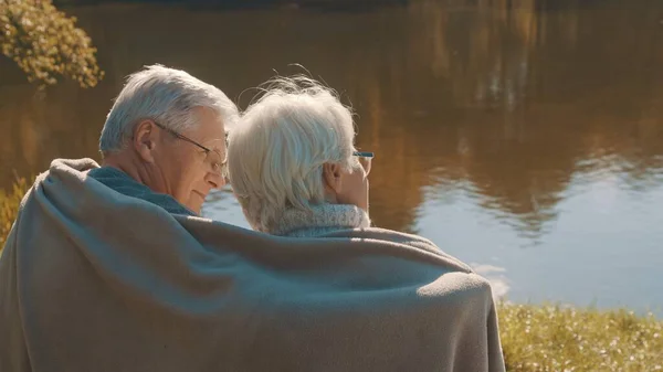 Pareja de ancianos de pelo gris sentados en el banco cerca del río cubriendo con manta el día de otoño. Romance y vejez —  Fotos de Stock