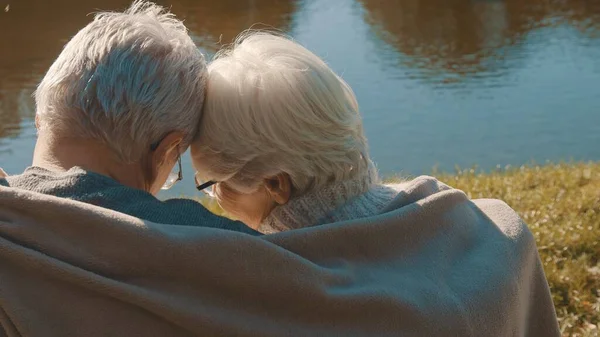 Ηλικιωμένο γκριζομάλλικο ζευγάρι κάθεται στον πάγκο κοντά στο ποτάμι καλύπτοντας με κουβέρτα την ημέρα του φθινοπώρου. Ειδύλλιο και γήρας — Φωτογραφία Αρχείου