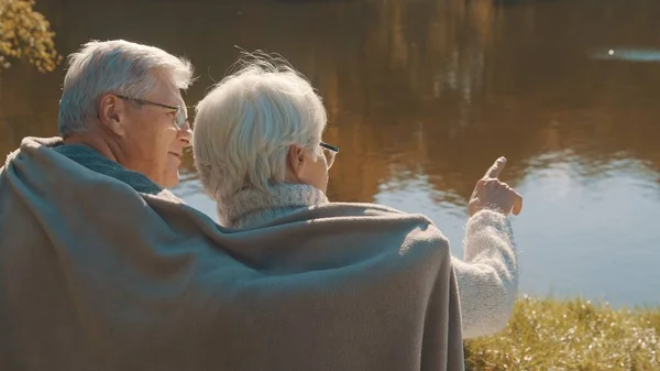 Pareja de ancianos de pelo gris sentados en el banco cerca del río cubriendo con manta el día de otoño. Romance y vejez —  Fotos de Stock