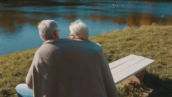 Ouder grijs harig echtpaar zittend op de bank in de buurt van de rivier bedekt met deken op herfstdag. Romantiek en ouderdom — Stockfoto