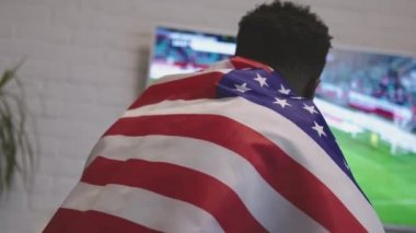 Heyecanlı siyah adam omuzlarının üzerinde Amerikan bayrağıyla akıllı televizyonda futbol maçı izliyor.