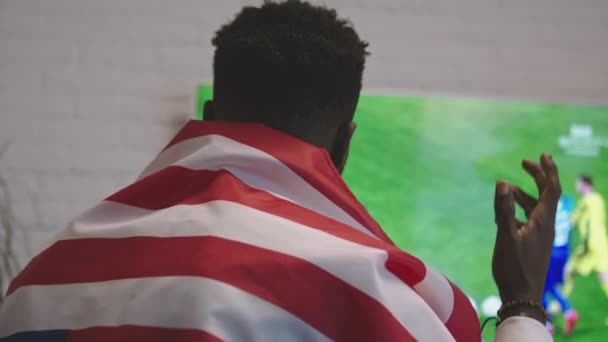 Homem negro desesperado com bandeira americana sobre os ombros assistindo jogo de futebol na TV inteligente. equipe favorita perder — Vídeo de Stock