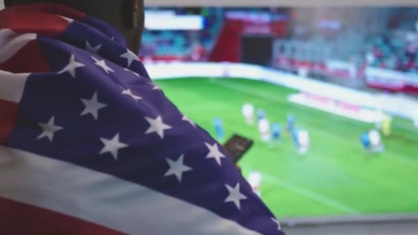 Hombre negro con bandera americana sobre los hombros viendo el partido de fútbol en la televisión inteligente — Vídeo de stock