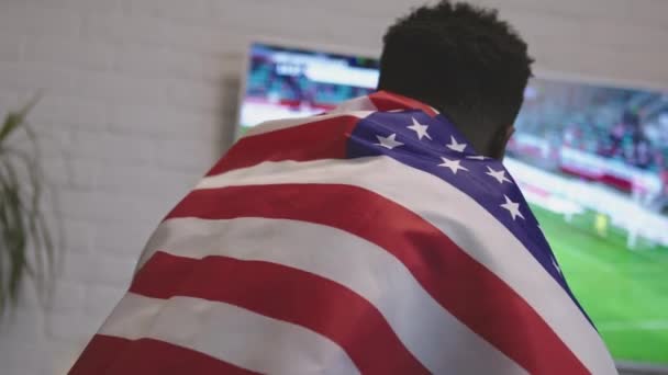 Opgewonden zwarte man met Amerikaanse vlag over de schouders kijken naar voetbalwedstrijd op smart tv — Stockvideo