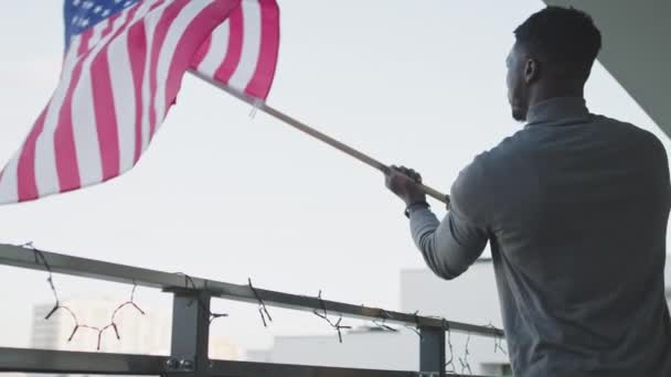 Junger attraktiver schwarzer Mann schwenkt amerikanische Flagge vom Balkon — Stockvideo