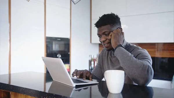 Trabajando remotamente. Africano americano negro hombre de negocios teniendo llamada telefónica en su apartamento mientras mira el portátil — Foto de Stock