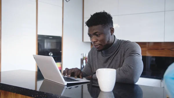 Genç Afrikalı Amerikalı siyah adam evinde dizüstü bilgisayarla çalışıyor ve çay ya da kahve içiyor. — Stok fotoğraf