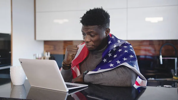 Homem afro-americano com bandeira dos EUA sobre seus ombros assistindo jogo em seu laptop com xícara de chá na mesa. Expressão facial surpresa — Fotografia de Stock