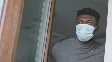Tıbbi maske takan siyah bir adam pencerenin yanında durmuş dışarıya bakıyor. Karantina ve zihinsel sağlık kavramı