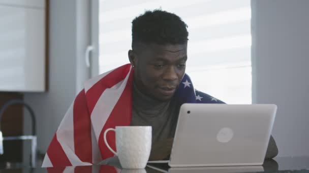 Un afro-american cu steag american deasupra umerilor se uită la joc pe laptop cu o ceașcă de ceai pe masă. Surprinsă expresie wace — Videoclip de stoc